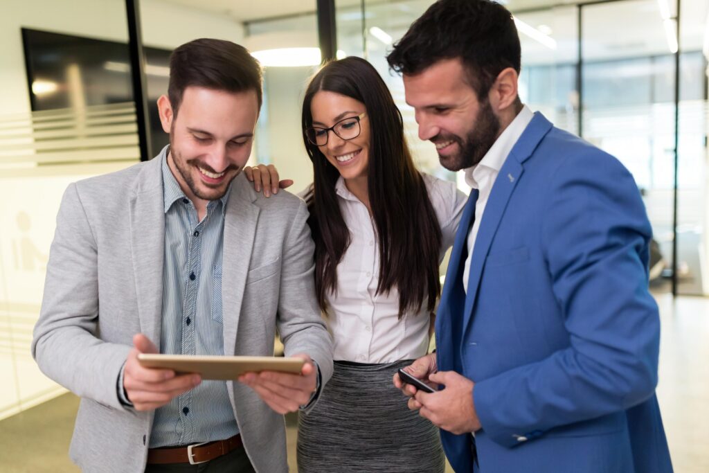 Three business professionals, two men and one woman, smiling and looking at a tablet together in a modern office environment, showcasing teamwork and collaboration