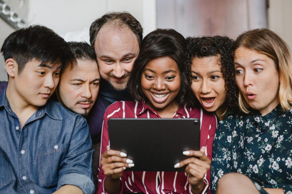 A diverse group of six colleagues are gathered closely together, all focused intently on a tablet screen held by one of them. Their expressions range from curiosity to excitement, indicating they are engaged and entertained by what they are viewing. The setting appears casual, suggesting a relaxed and informal moment of collaboration or enjoyment among the team. This image highlights teamwork, diversity, and the engaging nature of digital content.