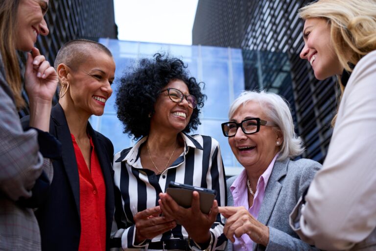 A group of diverse professional women smiling and engaging in a conversation while looking at a tablet, standing outside in a modern corporate environment.