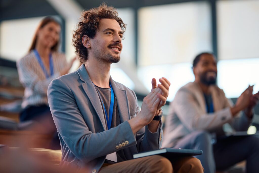 A young entrepreneur clapping during a successful business presentation at a conference, with other attendees in the background showing their appreciation.