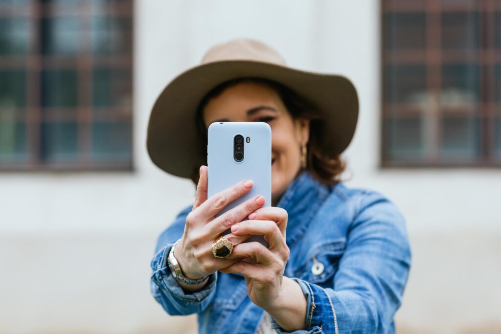 A woman wearing a denim jacket and a wide-brimmed hat is holding a smartphone in front of her, taking a selfie. Her face is partially obscured by the phone, but her smile and joy are evident. The background is slightly blurred, featuring a building with large windows. This image highlights individual exploration, enjoyment, and the use of mobile technology to capture moments, aligning with the spirit of MooveGO's self-guided tours and adventures.