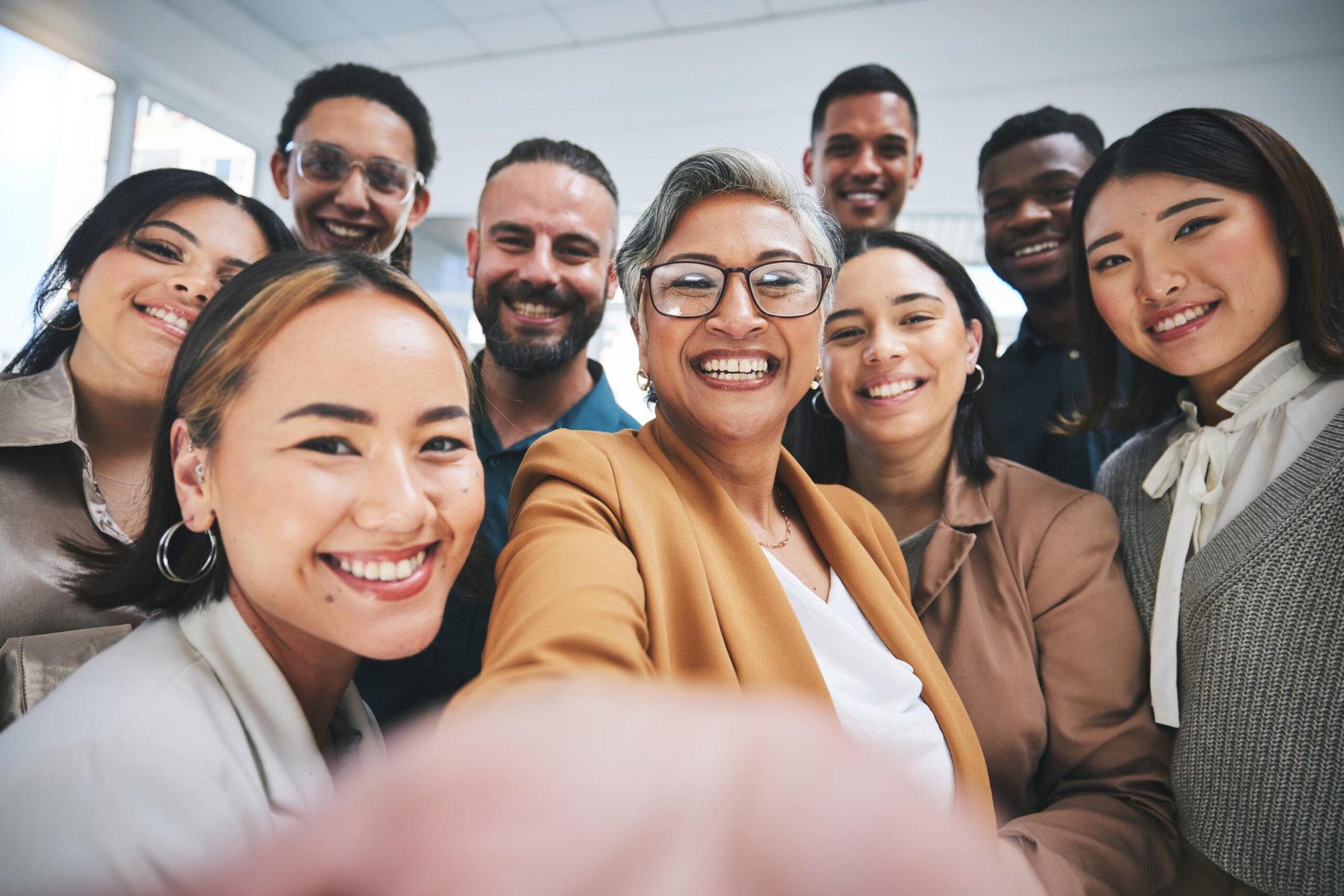 A group of diverse business colleagues smiling and taking a selfie together in a professional office setting, showcasing teamwork and positive corporate culture.