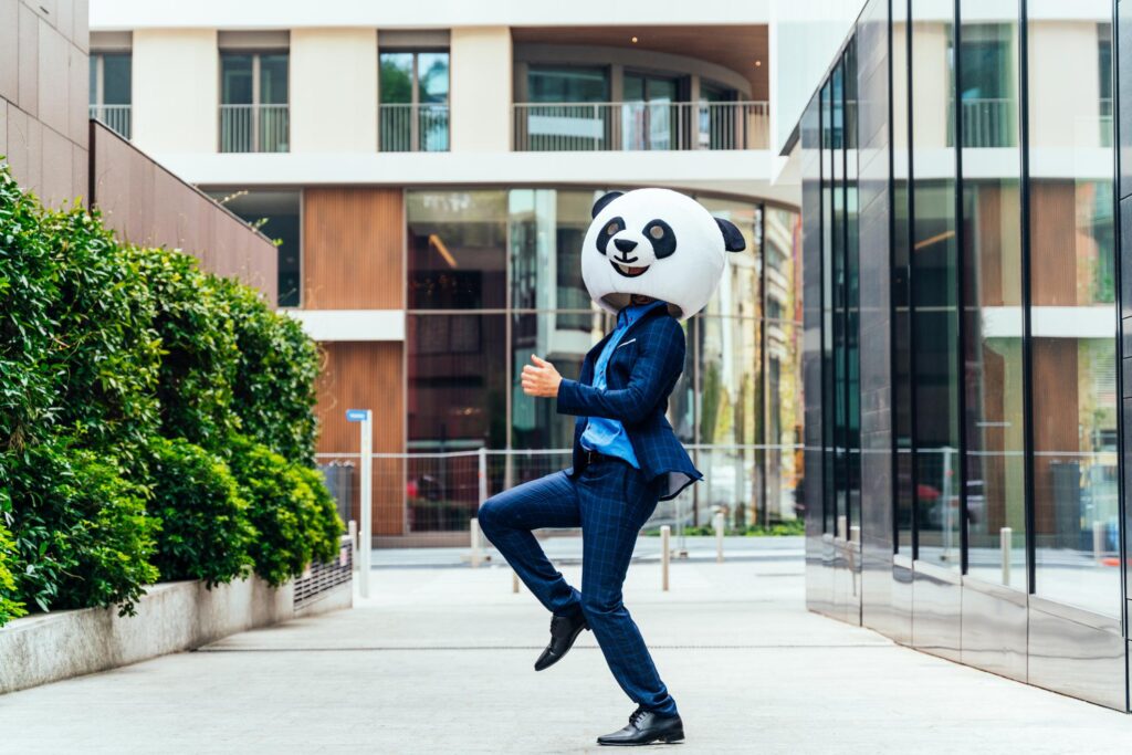 A businessman in a blue suit wearing a giant panda mask performs a playful dance in an urban setting, adding a fun and lighthearted element perfect for Halloween team-building activities.