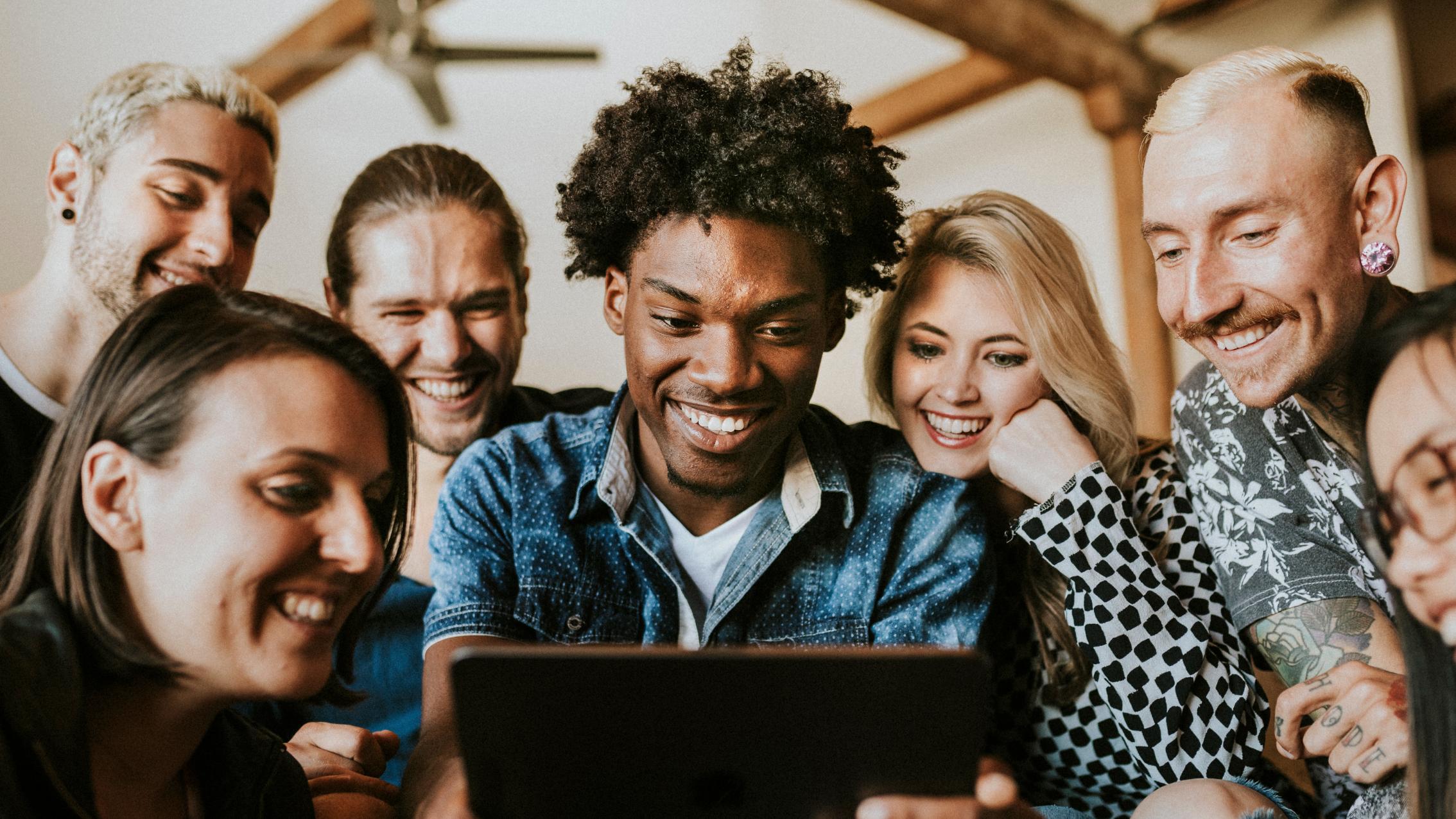 A group of diverse people smiling and gathered around a digital tablet, engaging together in a shared activity. The setting appears casual, and the atmosphere is joyful and collaborative.