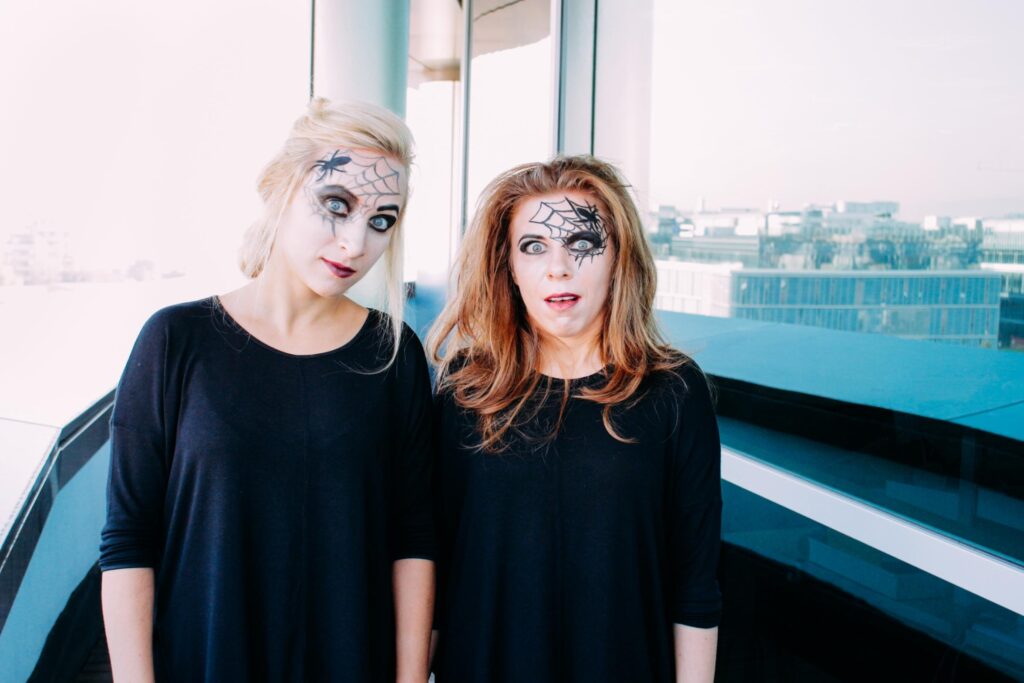 Two women with spider web face paint, standing in front of a large window in an office building. Both are wearing black outfits and have dramatic Halloween makeup, creating a spooky and fun atmosphere.