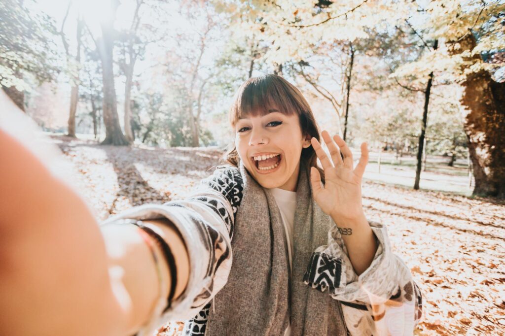 A young woman smiling and waving while taking a selfie in an autumn park. She is wearing a cozy scarf and sweater, with fallen leaves scattered on the ground and sunlight filtering through the trees in the background.