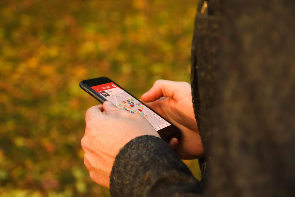 Close-up of a person holding a smartphone outdoors, displaying the Mooveteam app with a geolocation map on the screen. The background shows autumn leaves, creating a fall atmosphere, suggesting an outdoor team-building activity using Mooveteam's mobile technology.