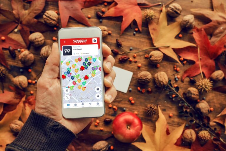 A hand holding a smartphone with the Mooveteam app open, displaying a geolocation map on the screen. The phone is set against a background of autumn decorations, including fallen leaves, walnuts, and an apple, creating a seasonal, fall-themed atmosphere.