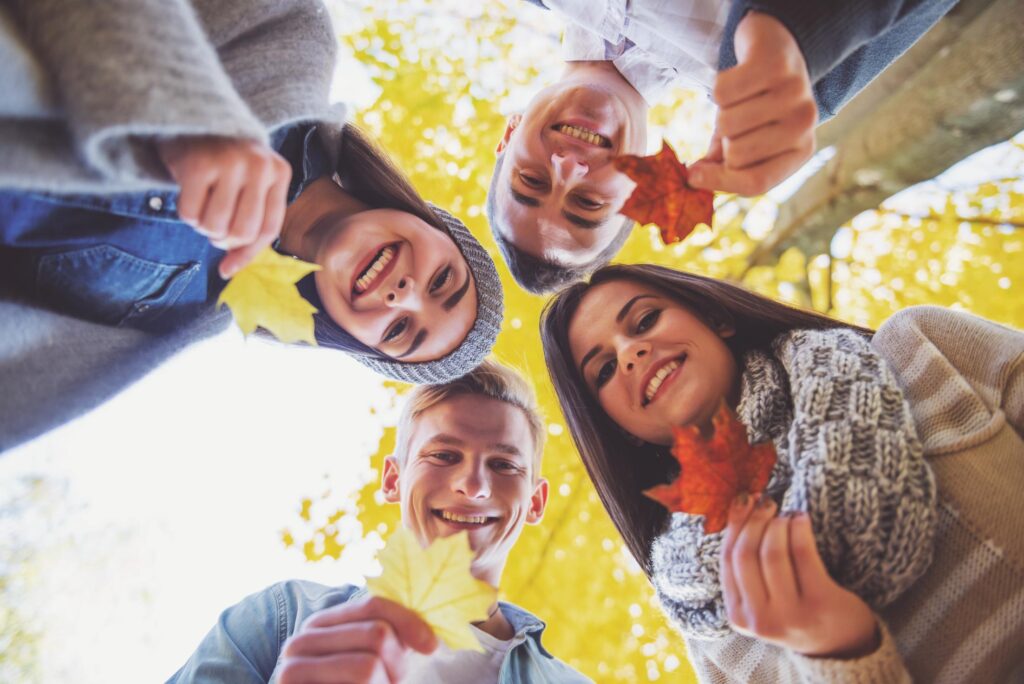 A group of four smiling young adults holding autumn leaves, looking down towards the camera in an outdoor setting with yellow and orange fall foliage in the background. They are dressed in cozy fall clothing, creating a warm, seasonal atmosphere.