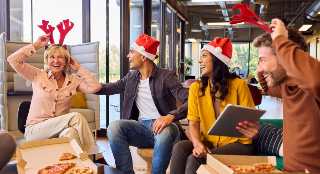 A group of four colleagues is enjoying a festive office Christmas party. They are wearing Santa hats and reindeer antlers, smiling and laughing together. One person is holding a tablet, and there are pizza boxes on the table, creating a relaxed and cheerful atmosphere. The background shows a bright, modern office space with large windows and natural light.