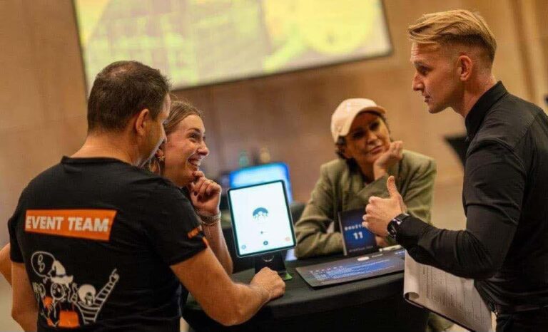 A dynamic team-building session with four participants engaged in a lively discussion around a tablet device. The group includes a facilitator wearing an "Event Team" shirt, with a vibrant and collaborative atmosphere in the background.