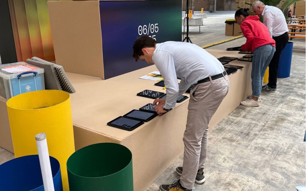 Dos personas colocan varias tablets en una mesa elevada junto a un cubo decorativo con la fecha ‘06/05/25’. Una mujer vestida con sudadera roja y un hombre con camisa de vestir están alineando los dispositivos antes de entregarlos. Al fondo, se ven más mesas numeradas y elementos logísticos del evento, todo bajo vigas y arcos de ladrillo