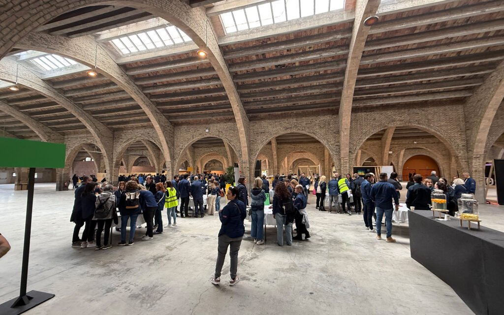 Gran sala con bóvedas de ladrillo y techo de vigas visto desde el fondo. Grupos de participantes se reúnen alrededor de varias mesas redondas, cada una identificada con un cartel numerado. La mayoría lleva chaquetas oscuras y se observa a una persona con chaleco reflectante coordinando. Al fondo, mesas adicionales y una estación de refrescos a la derecha, todo sobre un suelo de hormigón claro.