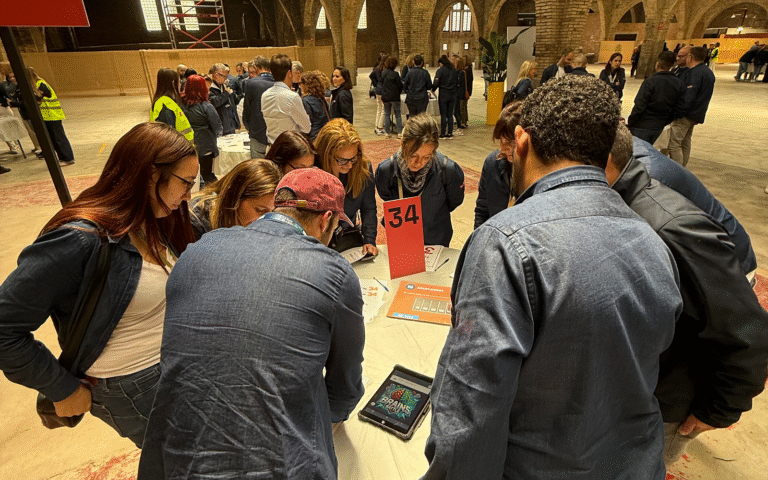 Un grupo de personas se reúne alrededor de una mesa redonda marcada con el cartel número 34. Sobre la mesa hay hojas naranjas con preguntas impresas (‘La Marató’ y otros retos) y una tablet que muestra la pantalla de inicio de ‘Brains Battle’. Los participantes, en su mayoría con chaquetas azules, están inclinados concentrándose en resolver el desafío, mientras en el fondo se ven más equipos y el amplio espacio del recinto con bóvedas de ladrillo.