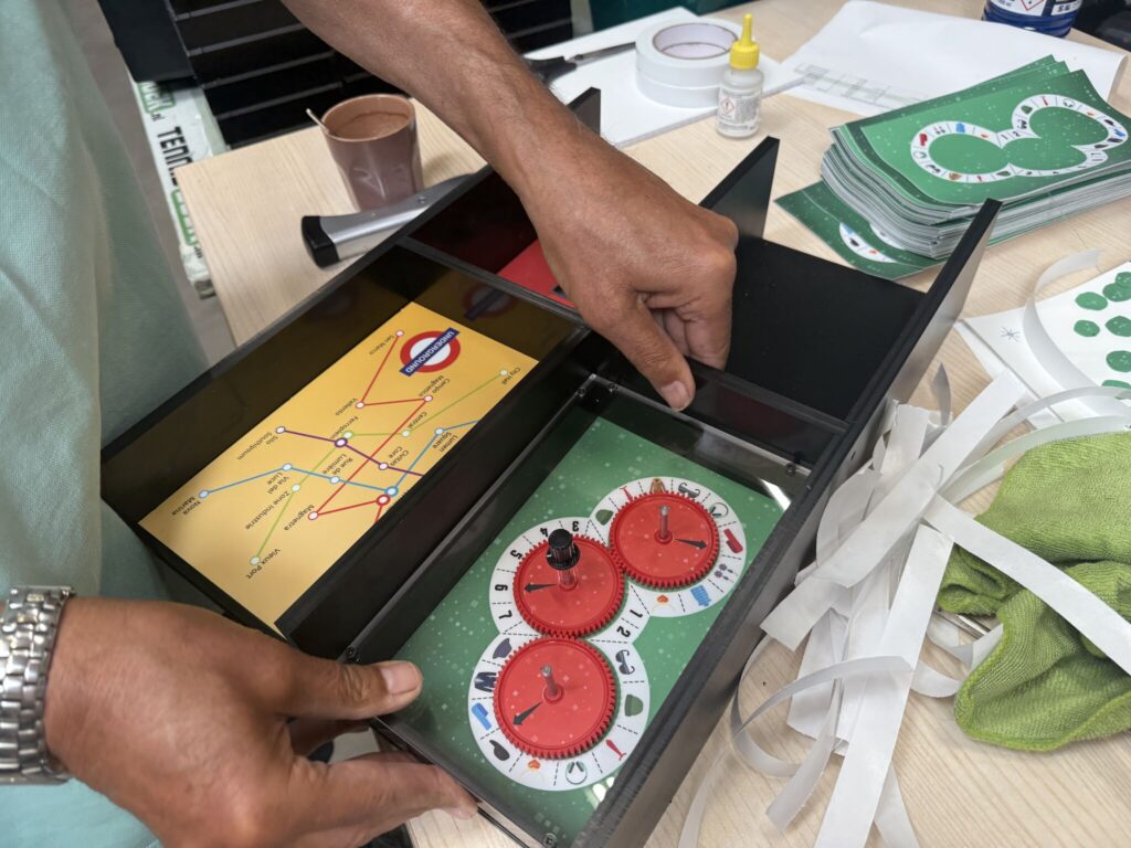 Close-up of a person assembling a Seconds2Survive EscapeBox, revealing internal puzzle components including red decoder dials and a map resembling a subway system. Surrounding the box are game cards, tape strips, and assembly tools on a work table.