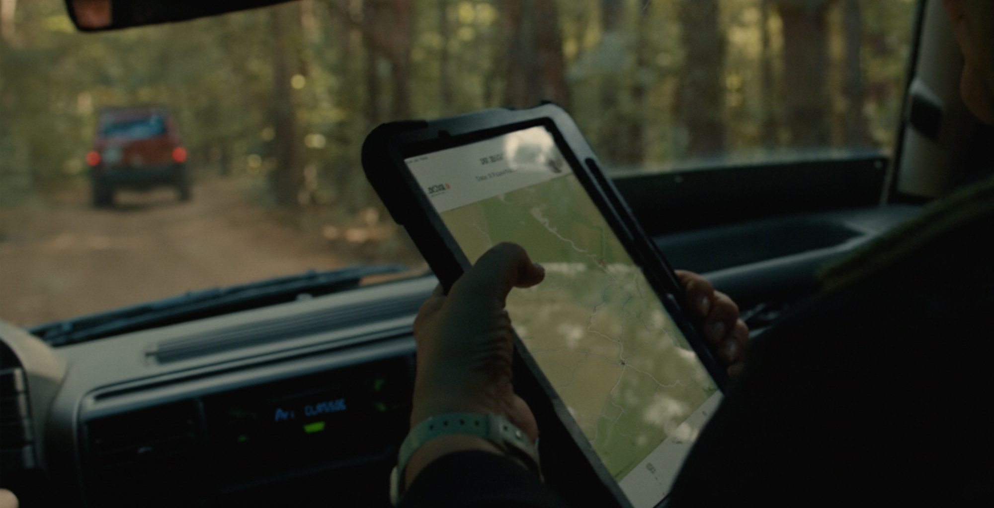 A person in a car holds a tablet displaying a map, following another vehicle on a dirt road.